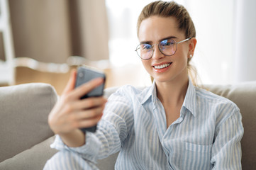 Amazing caucasian girl with a charming smile and with glasses takes a selfie on her phone at home