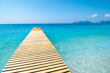 A wooden jetty extends into a calm and tranquil blue Mediterranean Sea on a sunny day with a clear blue sky.