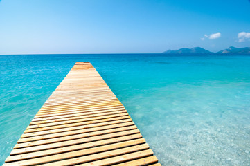 Fototapeta premium A wooden jetty extends into a calm and tranquil blue Mediterranean Sea on a sunny day with a clear blue sky.