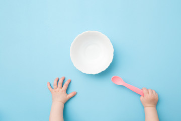Baby hands holding pink plastic spoon and waiting food. Empty white bowl on light blue table...