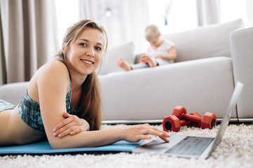Sport at home. Young attractive mom doing sports at home and watching a video fitness lesson on a laptop, and her little baby girl sit on a couch