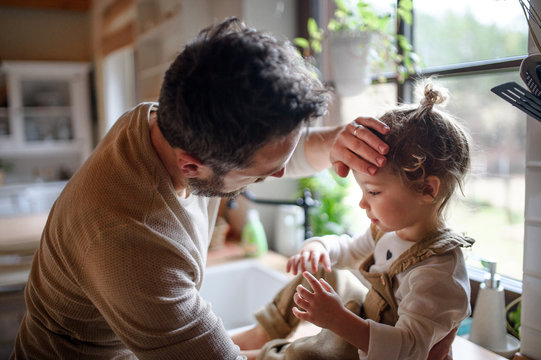 Father checking forehead of sick toddler daughter indoors at home.