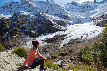 Travel woman sitting on the top and looking at the snowy mountains of Alibek glasier in sunny day in Dombay mountains, trekking in national park Russia