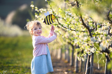 Small toddler girl standing outdoors in orchard in spring, holding paper bee.