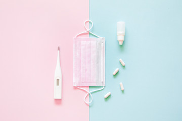 Top view of medical disposable mask, pills and a thermometer on pink and green flatlay background. Coronavirus disease or corona virus concept. Space for text.