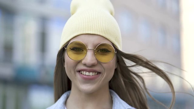4k. Real young smiling girl in yellow cap and glasses stand in city area. Street portrait

