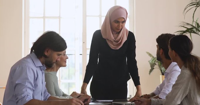 Pleasant young asian islamic female team leader in hijab standing at table, holding business meeting with diverse colleagues. Concentrated multicultural team with arab boss discussing financial data.