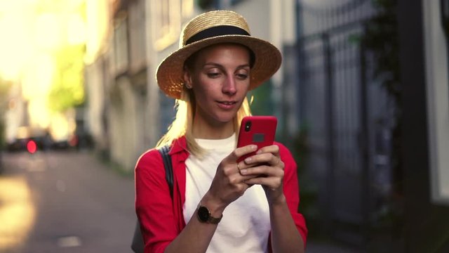 
Slow motion effect of cheerful woman in hat using mobile phone browsing network while walking outdoors on city street. Positive caucasian hipster girl connected to 4g wireless internet connection 
