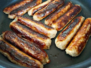 A close up view of fresh British Cumberland pork sausages sizzling in a frying pan. Cooking and preparing a sausage sandwich. Fatty and tasty food. Traditional English meat breakfast in a skillet pan.