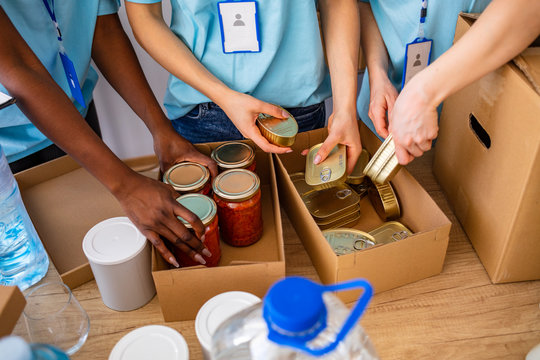 Homeless care. Charity team of volunteers putting food and drinks into paper box. Volunteers Collecting Food Donations In Warehouse. Volunteers with donation box with foodstuffs on wooden background