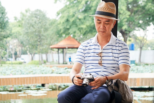 Elderly Chinese man resting by pond in park and checking photos he made on new digital camera during walk