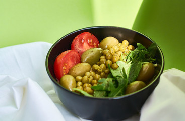 Green summer background and white cloth with  black bowl and delicious useful breakfast of porridge and vegetables. Tomatoes, olives, arugula, peas.