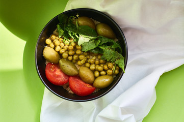 Green summer background and white cloth with  black bowl and delicious useful breakfast of porridge and vegetables. Tomatoes, olives, arugula, peas.