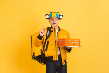 Holding pizza, pointing up. Emotions of caucasian deliveryman isolated on yellow background. Contacless delivery service during quarantine. Man delivers food during isolation. Safety. Hurrying up.