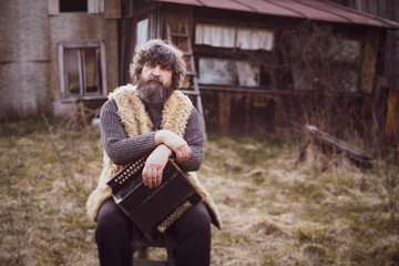 curly haired adult man with a beard, sitting with an old accordion in the background of his...