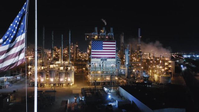 American flag waving on the wind against the background of petroleum refinery. Night aerial shot. Plant facilities are illuminated in the dark. Another star-striped flag is on metal construction. 4K
