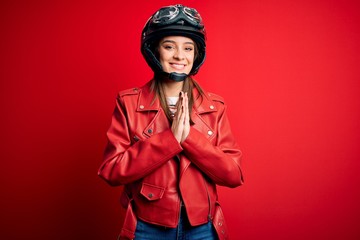 Young beautiful brunette motocyclist woman wearing motorcycle helmet and red jacket praying with...