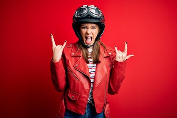 Young beautiful brunette motocyclist woman wearing motorcycle helmet and red jacket shouting with crazy expression doing rock symbol with hands up. Music star. Heavy concept.