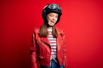 Young beautiful brunette motocyclist woman wearing motorcycle helmet and red jacket winking looking at the camera with sexy expression, cheerful and happy face.