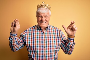 Senior handsome hoary man wearing golden crown of king over isolated yellow background gesturing finger crossed smiling with hope and eyes closed. Luck and superstitious concept.