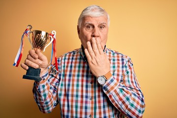 Senior handsome hoary champion man holding trophy award for victory over yellow background cover mouth with hand shocked with shame for mistake, expression of fear, scared in silence, secret concept