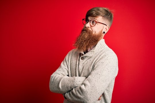 Handsome Irish redhead man with beard wearing casual sweater and glasses over red background looking to the side with arms crossed convinced and confident
