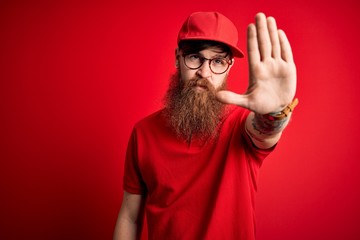 Young handsome delivery man wearing glasses and red cap over isolated background doing stop sing with palm of the hand. Warning expression with negative and serious gesture on the face.