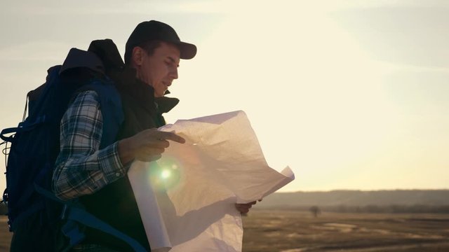 A man, a tourist with a backpack is standing in an open area and reading a map of the location.