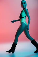 Fototapeta na wymiar fashionable young model in futuristic leotard and fire-shaped sunglasses posing on pink in blue light