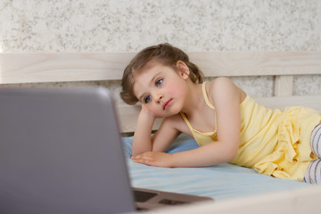 girl 3 - 4 years Caucasian in yellow dress lying on wooden bed on blue bedspread and looking at laptop, concept of home education