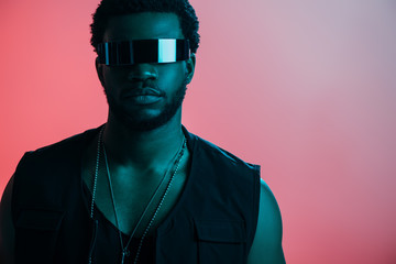 young stylish african american man in futuristic sunglasses posing on pink in blue light