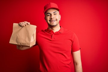 Young handsome delivery man holding paper bag with takeaway food over red background with a happy face standing and smiling with a confident smile showing teeth