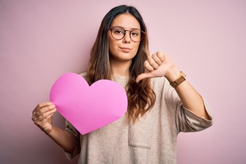 Young beautiful brunette romantic woman holding big heart paper celebrating valentine day with angry face, negative sign showing dislike with thumbs down, rejection concept