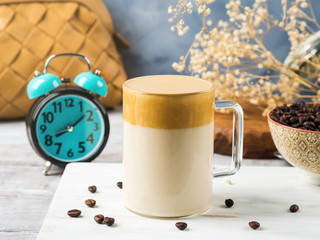 Obraz na płótnie Canvas Whipped dalgona coffee drink in glass mug on white wooden board. Cozy breakfast with trendy beverage. Alarm clock