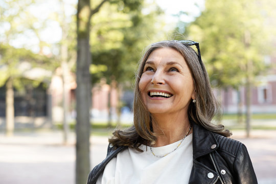 Cheerful overjoyed middle aged woman wearing sunglasses on her head and black leather jacket over white blouse enjoying peaceful beautiful morning while waking in park, looking up with broad smile