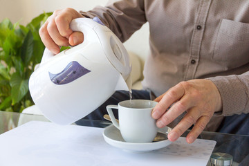 Close-up hands of a senior man. A pensioner pours water from a white kettle into a coffee cup.