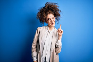 Young beautiful businesswoman with curly hair and piercing wearing jacket and glasses pointing finger up with successful idea. Exited and happy. Number one.