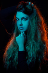 fashion portrait of a blonde girl with color gel filters, neon skin. she holds her hand under her chin, with her second hand holds her hair. blue face, orange hair in the back