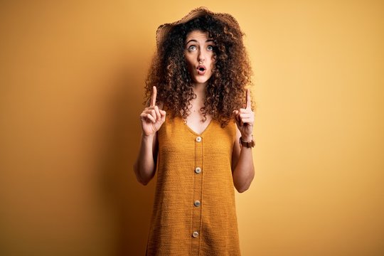Beautiful brunette woman on vacation with curly hair and piercing wearing hat and dress amazed and surprised looking up and pointing with fingers and raised arms.