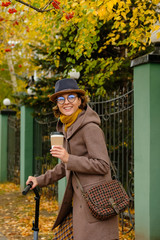 Beautiful girl in a hat and glasses riding a scooter on the background of autumn. Modern young woman drinks coffee in a paper cup on her way to work.