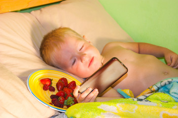 Morning at the cottage - a little boy with blond hair and blue eyes lies on the bed, happily squinting at the sunlight and watching cartoons in the phone.