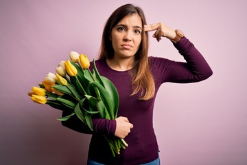 Young blonde woman holding romantic bouquet of yellow tulips flowers over pink background Shooting and killing oneself pointing hand and fingers to head like gun, suicide gesture.