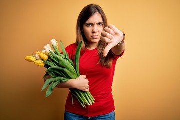 Young blonde woman holding romantic bouquet of tulips flowers over yellow background looking unhappy and angry showing rejection and negative with thumbs down gesture. Bad expression.