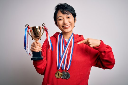 Young beautiful asian girl winner holding trophy wearing medals over white background with surprise face pointing finger to himself