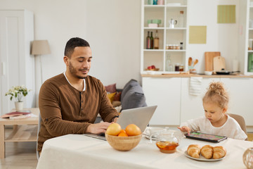 Fototapeta na wymiar Young man using laptop, his little daughter sitting at table next to him playing game on digital tablet, horizontal shot