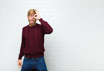 young blonde man smiling happily with funny face, joking and looking through peephole, spying on...