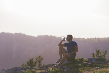 Handsome athlete sitting at a rocky peak while looking at the breathtaking mountain line and a beautiful lake while wearing a blue shirt and grey shorts.