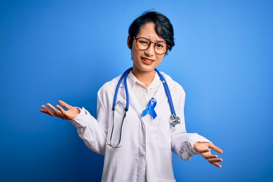 Young beautiful asian doctor girl wearing stethoscope and coat with blue cancer ribbon clueless and confused expression with arms and hands raised. Doubt concept.