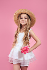Stylish kid girl in straw hat and white dress posing over pink background