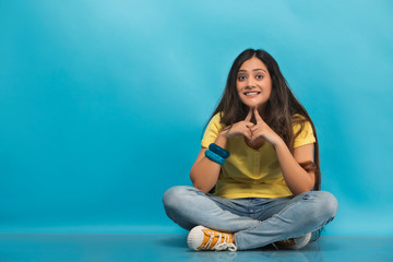 Smiling teenage girl in jeans and t-shirt sitting cross legged on the floor with her hands touching her chin
